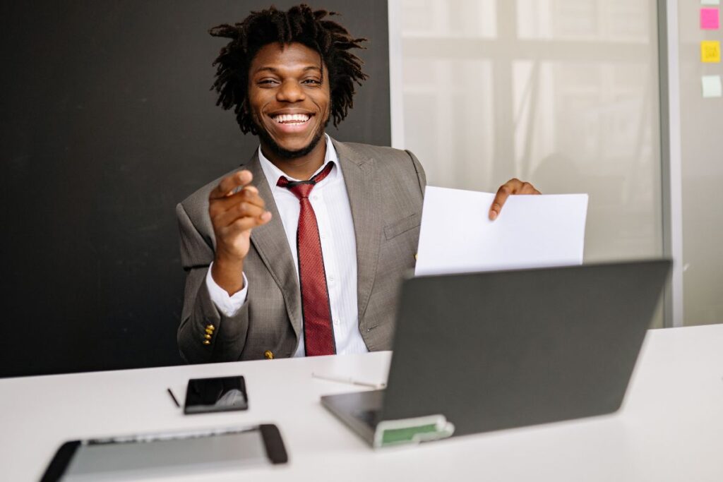 Um jovem profissional sorridente em traje social aponta para a câmera enquanto segura um documento, exemplificando a confiança necessária para conseguir emprego em 2026 e as novas oportunidades de carreira.